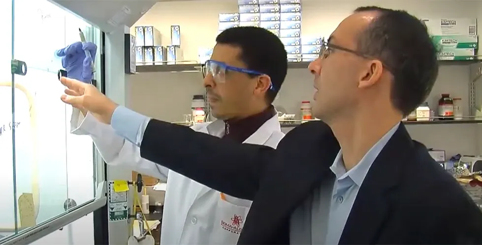 Dr. Brent Stockwell and a researcher discuss data at a laboratory workstation, pointing to a diagram on a glass panel. 