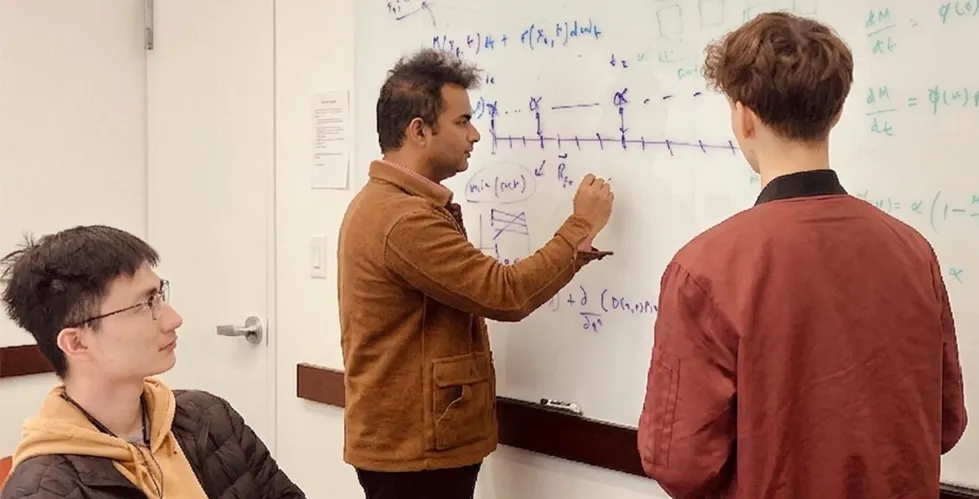Dr. Sanket Rane writes equations on a whiteboard while conversing with two members of his lab group.