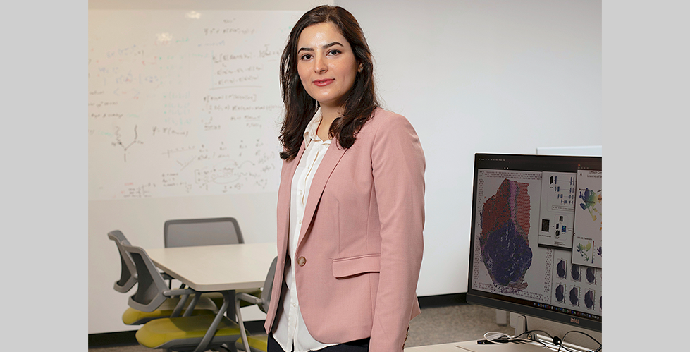 Photography of Elham Azizi in front of a white boards displaying chemical structures and in front of a computer